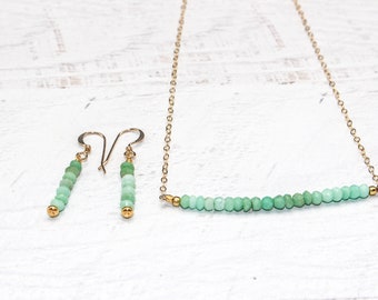 Op de afbeelding: Een delicate sieradenset met een ketting en oorbellen. De ketting en oorbellen hebben een gouden ketting en haken, met een verloop van groene kralen. De oorbellen zijn klein en de ketting is een horizontale balk.