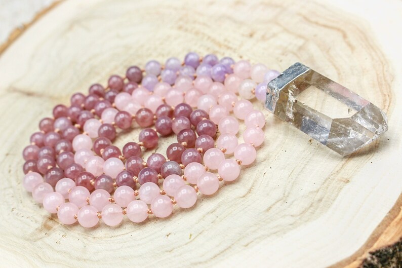 May include: A beaded mala necklace with a gradient of pink and purple beads, alongside a clear quartz crystal. The beads transition from a light pink to a deeper purple. The necklace is displayed on a wooden surface.