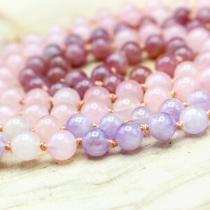 May include: A beaded necklace featuring a gradient of pink and purple gemstones. The beads range in color from light pink to deep purple. The necklace is strung on a light brown cord, and the beads are round and polished.