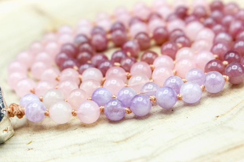 May include: A beaded necklace featuring a gradient of pink and purple gemstones. The beads range in color from light pink to deep purple. The necklace is strung on a light brown cord, and the beads are round and polished.