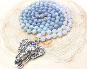 May include: A beaded necklace with a silver elephant pendant. The necklace features light blue and white beads. The elephant pendant has intricate details and a blue eye. The necklace is displayed on a wooden surface.