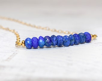 May include: A delicate gold chain necklace featuring a row of faceted, deep blue lapis lazuli beads. The beads are strung horizontally, creating a bar shape, with gold accents at each end. The necklace is displayed on a light-colored surface.