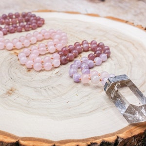 May include: Three beaded necklaces in shades of pink and purple, with a clear crystal pendant, rest on a wooden surface. The necklaces are made of small, round beads strung together. The crystal is rectangular with a silver cap.