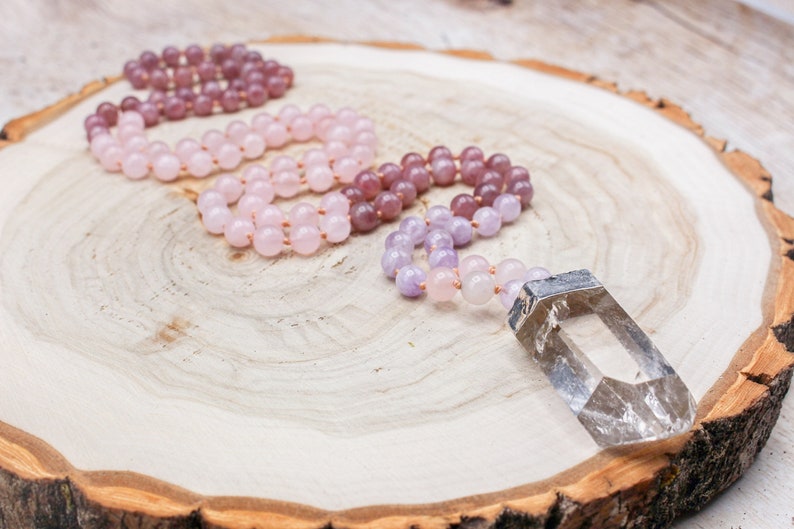 May include: Three beaded necklaces in shades of pink and purple, with a clear crystal pendant, rest on a wooden surface. The necklaces are made of small, round beads strung together. The crystal is rectangular with a silver cap.