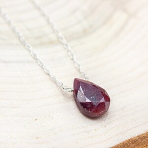 Op de afbeelding: Een zilveren ketting met een druppelvormige, gefacetteerde robijn edelsteen hanger. De robijn heeft een dieprode kleur en hangt aan een fijne zilveren ketting. De ketting wordt gepresenteerd op een licht houten oppervlak.