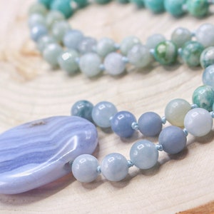 May include: A necklace featuring a large, smooth, oval-shaped blue lace agate pendant with distinct white banding, strung on a cord of round, polished beads in varying shades of light blue, teal, and soft green, knotted between each bead.