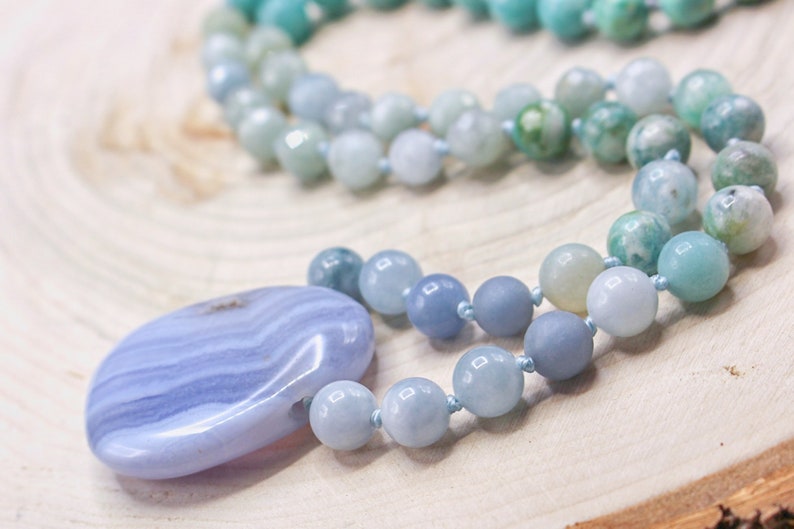 May include: A necklace featuring a large, smooth, oval-shaped blue lace agate pendant with distinct white banding, strung on a cord of round, polished beads in varying shades of light blue, teal, and soft green, knotted between each bead.