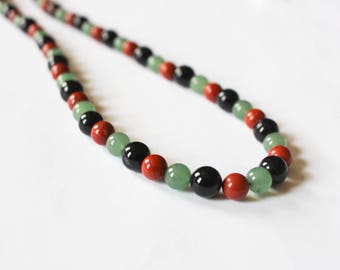 May include: A beaded necklace featuring a pattern of black, red, and green beads. The beads are round and appear to be polished. The necklace is displayed against a white background.