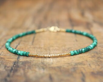 May include: A delicate bracelet featuring small, faceted, emerald green beads interspersed with a section of gold-colored, rectangular beads. The bracelet is strung on a thin, gold-colored chain with a clasp.