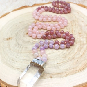 May include: Three gemstone malas in shades of pink and purple rest on a wooden surface. The malas are made of small, round beads. One mala has a clear quartz crystal pendant with a silver-colored metal cap.