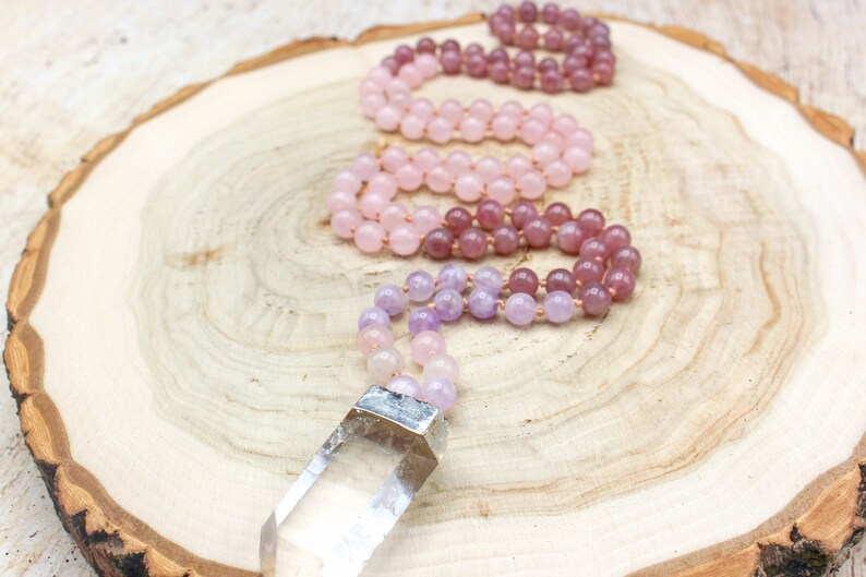 May include: Three gemstone malas in shades of pink and purple rest on a wooden surface. The malas are made of small, round beads. One mala has a clear quartz crystal pendant with a silver-colored metal cap.