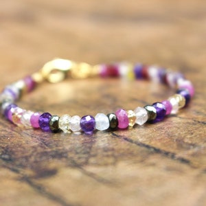 May include: A beaded bracelet featuring a variety of colorful gemstones. The bracelet includes purple, pink, clear, and gold-colored beads. The clasp is gold-toned. The bracelet is displayed on a wooden surface.