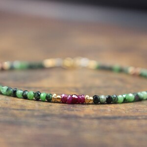 May include: A delicate beaded bracelet featuring green, black, and ruby-colored gemstones. The bracelet has gold-colored accents and a clasp. The gemstones are small and round, strung together to form a flexible band.