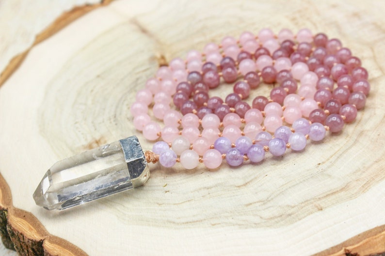 May include: A layered mala necklace featuring rose quartz, amethyst, and other pink and purple beads. The necklace is accented with a clear crystal point pendant with silver-colored metal accents. The beads are strung on a light brown cord.