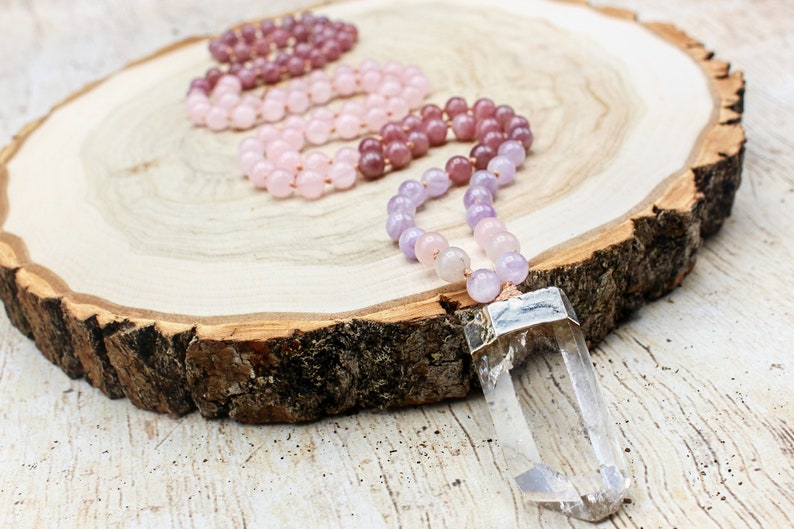 May include: A collection of beaded necklaces in shades of pink, purple, and clear quartz crystal pendants. The necklaces are displayed on a rustic wooden surface, showcasing the natural colors and textures of the materials.