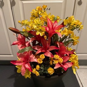 May include: A potted arrangement of pink and yellow artificial flowers. The flowers are in a black plastic pot and are sitting on a black mat.