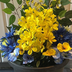 May include: A black planter filled with a floral arrangement of yellow and blue flowers. The flowers are arranged in a circular pattern with the yellow flowers in the center and the blue flowers around the edges.