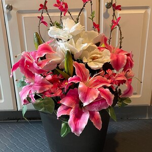 May include: A black planter with pink and white flowers. The flowers are lilies and magnolias. The planter is sitting on a gray tiled floor.