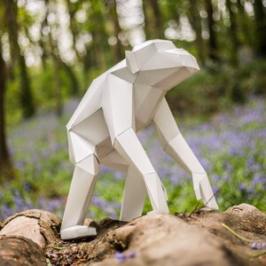 Peut inclure: Une sculpture en origami blanche d'un chimpanzé debout sur un tronc d'arbre dans un décor de forêt. La sculpture est en papier et a un design géométrique.