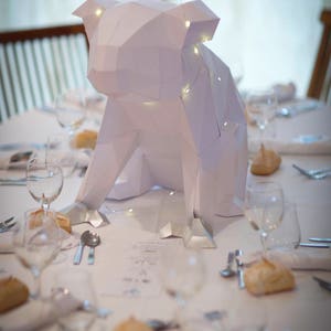 May include: A white paper origami animal sculpture of a dog with string lights inside, sitting on a white tablecloth with glasses and silverware on a table.