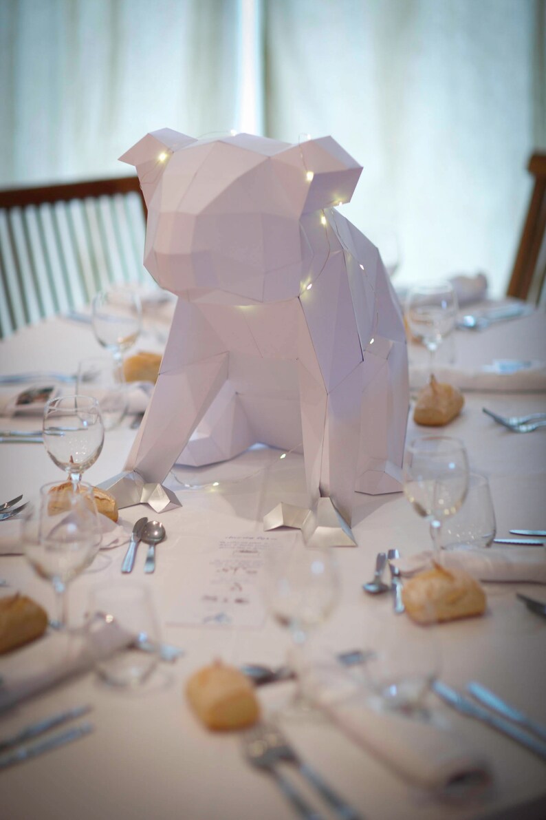 May include: A white paper origami animal sculpture of a dog with string lights inside, sitting on a white tablecloth with glasses and silverware on a table.