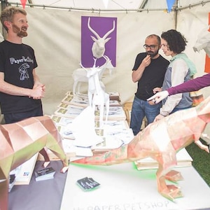 Puede incluir: Una tienda de artesan&iacute;a de papel con una variedad de animales de origami, incluyendo un T-Rex, un perro y varios ciervos. El due&ntilde;o de la tienda est&aacute; hablando con los clientes sobre la artesan&iacute;a de papel.