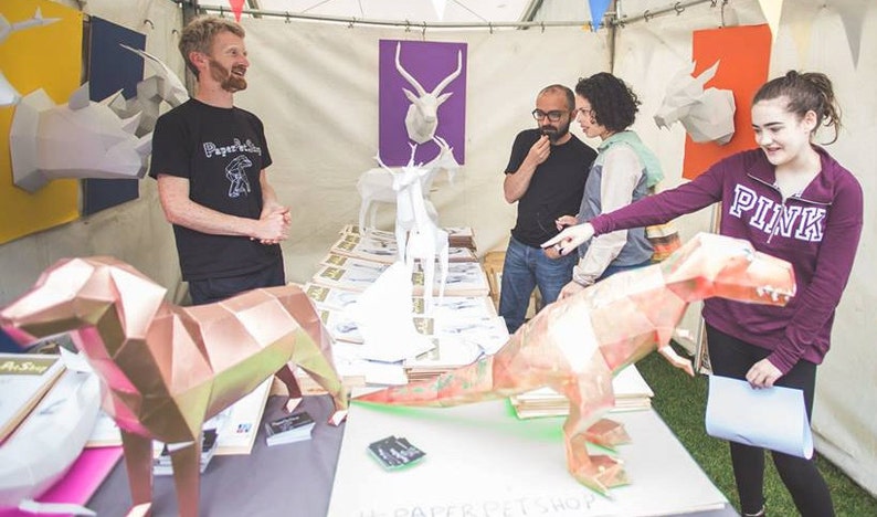 Puede incluir: Una tienda de artesan&iacute;a de papel con una variedad de animales de origami, incluyendo un T-Rex, un perro y varios ciervos. El due&ntilde;o de la tienda est&aacute; hablando con los clientes sobre la artesan&iacute;a de papel.