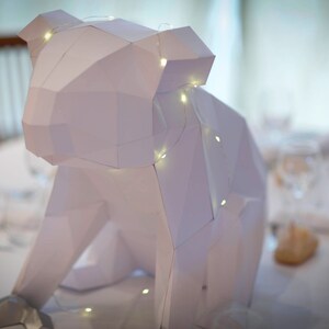 May include: A white paper sculpture of an animal, possibly a bear or a dog, with a string of lights wrapped around it. The sculpture is sitting on a white table with a glass of water and a napkin.