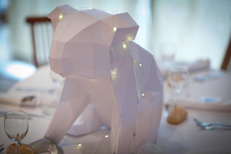 May include: A white paper sculpture of an animal, possibly a bear or a dog, with a string of lights wrapped around it. The sculpture is sitting on a white table with a glass of water and a napkin.