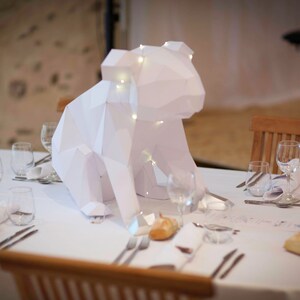 May include: A white origami animal sculpture, possibly a bear, sits on a white tablecloth with a place setting. The sculpture is lit with small white lights.
