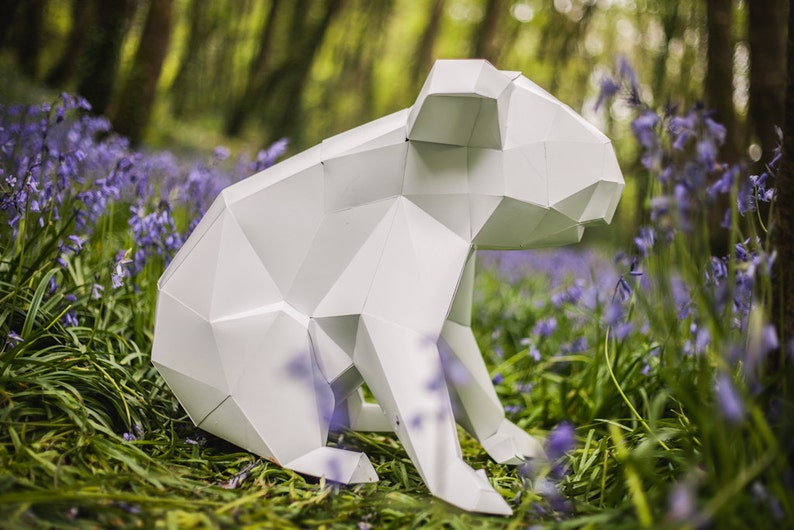 May include: A white paper origami koala sculpture sitting in a field of purple flowers.