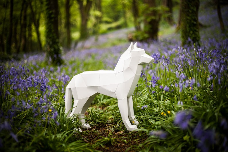 May include: A white paper wolf sculpture standing in a field of purple flowers. The sculpture is made of folded paper and has a realistic appearance.