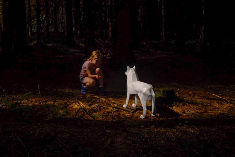 May include: A young person in a forest setting looking at a white paper wolf sculpture.