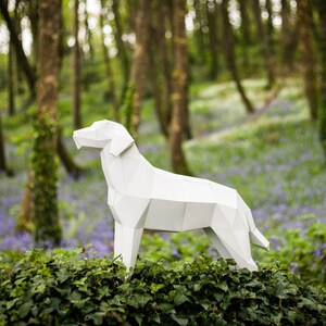 Puede incluir: Un modelo de papel blanco de un perro de pie en un entorno de bosque. El perro est&aacute; hecho de papel doblado y tiene una apariencia realista. El bosque est&aacute; lleno de flores azules.