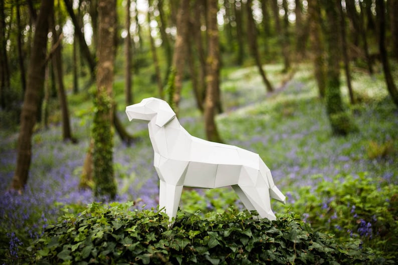 Puede incluir: Un modelo de papel blanco de un perro de pie en un entorno de bosque. El perro est&aacute; hecho de papel doblado y tiene una apariencia realista. El bosque est&aacute; lleno de flores azules.
