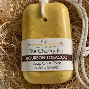 May include: A rectangular, yellow soap bar with a white label that reads "BOURBON TOBACCO Soap On A Rope HONEY & TURMERIC." The soap is attached to a white rope. The soap is in a bed of straw.