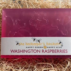 May include: A pink soap bar with a label that reads "the beekeeper's basket, happy queen, happy hive, WASHINGTON RASPBERRIES". The soap bar has a design of bees and flowers.