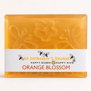 May include: A rectangular orange translucent soap bar featuring embossed bee and flower designs. A white paper band wraps around the center with the text The beekeeper's basket, HAPPY QUEEN, HAPPY HIVE, and ORANGE BLOSSOM printed in black and orange.