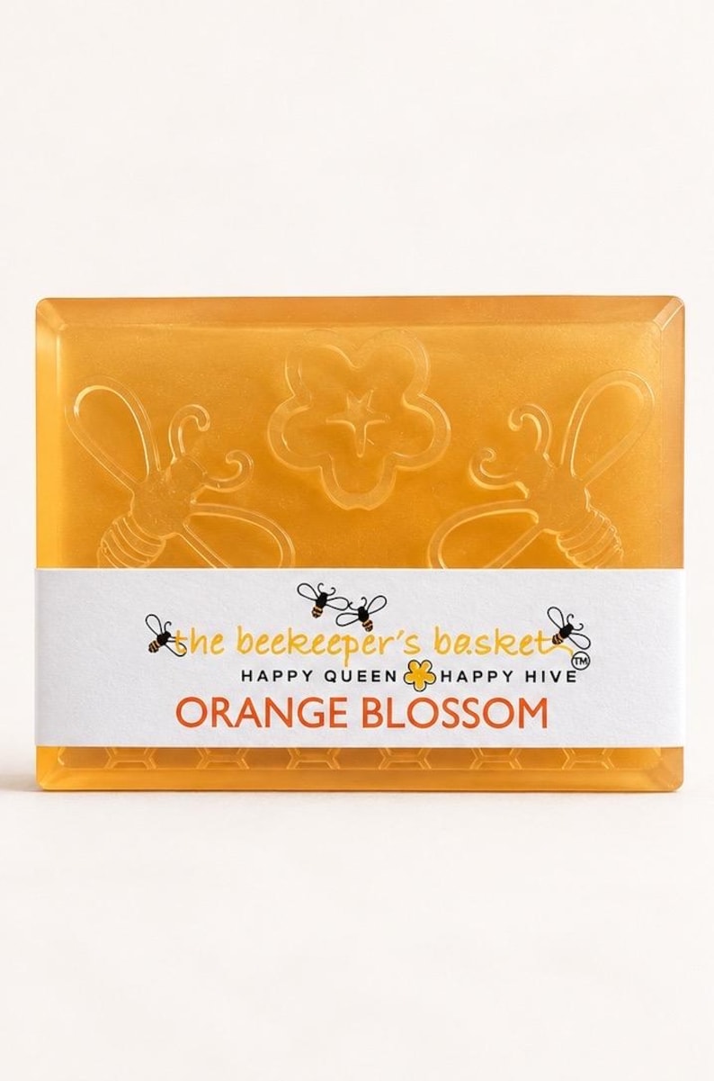 May include: A rectangular orange translucent soap bar featuring embossed bee and flower designs. A white paper band wraps around the center with the text The beekeeper's basket, HAPPY QUEEN, HAPPY HIVE, and ORANGE BLOSSOM printed in black and orange.