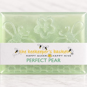 May include: A rectangular light green translucent soap bar featuring embossed bee and flower designs. A white paper band wraps around the center, printed with the text the beekeeper's basket, HAPPY QUEEN HAPPY HIVE, and PERFECT PEAR.