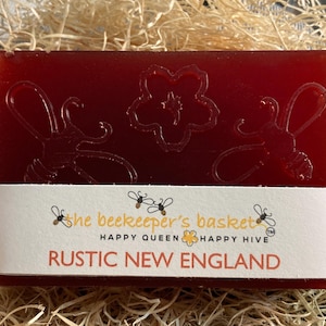 May include: A red bar of soap with a floral and bee design. The soap is labeled "the beekeeper's basket" and "RUSTIC NEW ENGLAND".