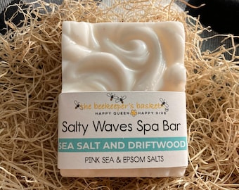 May include: A white spa bar with a swirl design, labeled "Salty Waves Spa Bar", "Sea Salt and Driftwood", and "Pink Sea & Epsom Salts". The bar is packaged in a clear plastic wrap and is sitting on a bed of shredded paper.