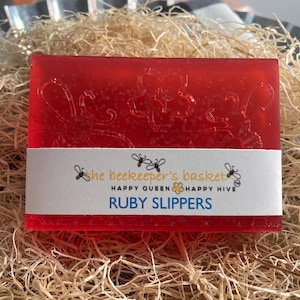 May include: A red soap bar with a white label that reads "The Beekeeper's Basket, Happy Queen, Happy Hive, Ruby Slippers".