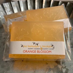 May include: Orange blossom scented soap bars in clear packaging. The bars are a light orange colour with floral designs. The label reads "Orange Blossom" with bee graphics and the text "Happy Queen Happy Hive".