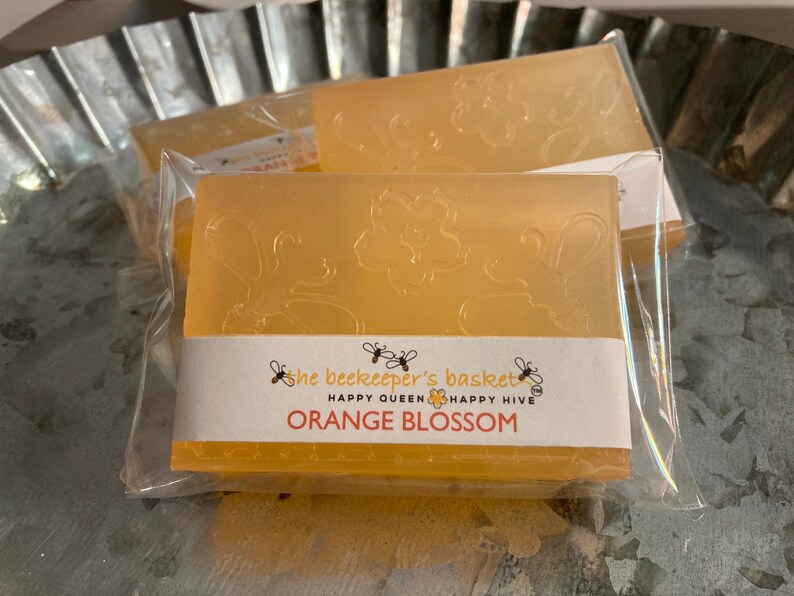 May include: Orange blossom scented soap bars in clear packaging. The bars are a light orange colour with floral designs. The label reads "Orange Blossom" with bee graphics and the text "Happy Queen Happy Hive".