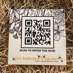 May include: A square, cream-coloured card with a large black and white QR code. The card features floral illustrations and the text "SCAN TO ENTER THE HIVE!" and "the beekeeper's basket" with bee graphics.