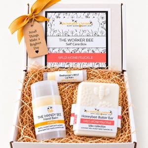 May include: A white gift box containing a Honeybee Butter Bar, a Handy Bee hand balm, and a Beekeeper's BEST lip balm. The box features a label reading The Worker Bee Self Care Box, Wild Honeysuckle, and a tag stating Small Things Make a Brighter Day.