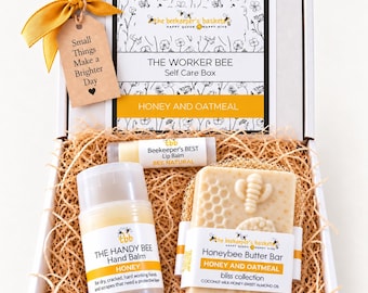 May include: A white gift box containing a Honey and Oatmeal self-care set. Items include a Honey hand balm stick, a Honeybee butter bar soap with a bee and honeycomb design, and a lip balm. A yellow ribbon and a tag reading Small Things Make a Brighter Day are attached.