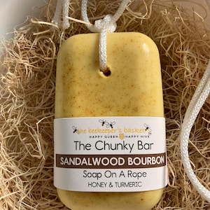 May include: A yellow bar of soap with a white rope attached. The soap is labeled "The Chunky Bar Sandalwood Bourbon Soap On A Rope Honey & Turmeric".