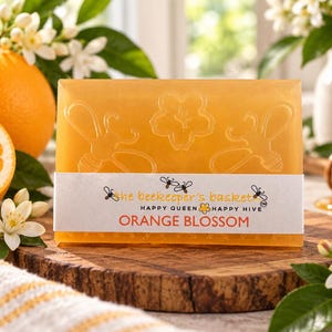 May include: A rectangular orange soap bar embossed with two bees and a flower. A white paper label wraps around the center, printed with the text "the beekeeper's basket", "HAPPY QUEEN HAPPY HIVE", and "ORANGE BLOSSOM" in orange and black ink.